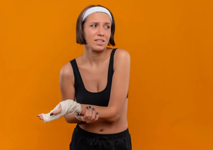 A woman with short brown hair and a white headband stands against an orange background. She looks concerned and holds her bandaged right hand with her left hand. She is wearing a black sports bra and shorts.