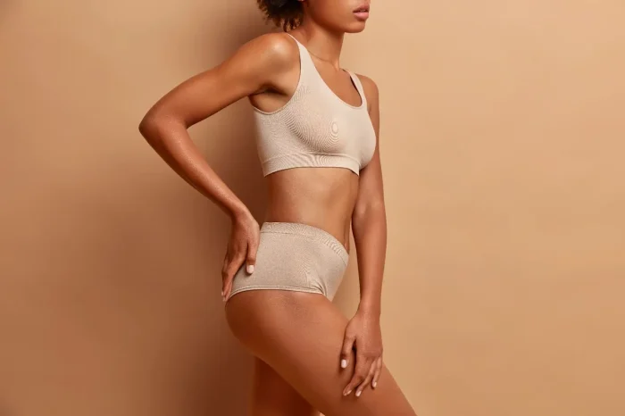 A woman poses in beige seamless underwear against a matching beige background, with one hand on her hip and her body turned to the side.