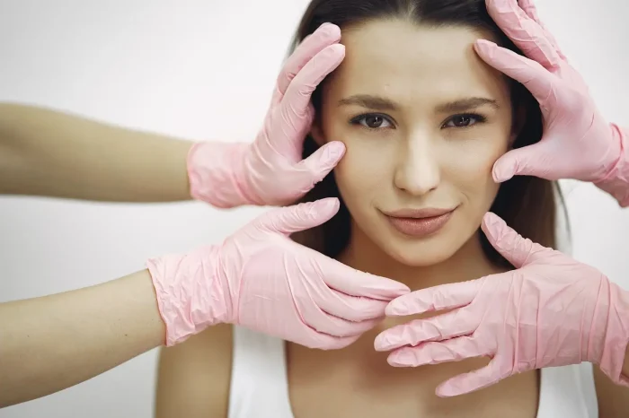 A woman with long brown hair is framed by several hands wearing pink gloves, gently touching her face. She looks directly at the camera with a neutral expression, suggesting a skincare or cosmetic procedure setting.
