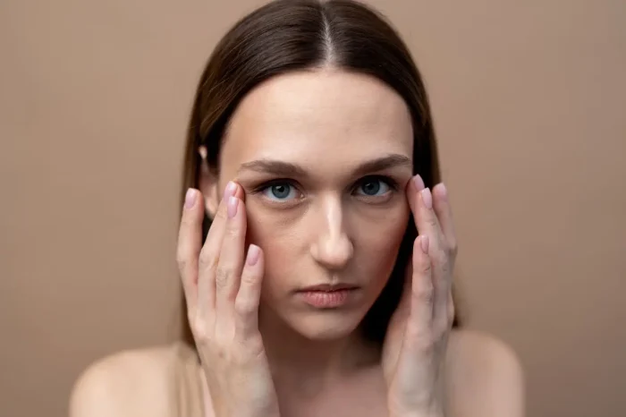 A woman with straight brown hair touches the sides of her face with both hands, looking directly at the camera against a plain beige background. She has a neutral expression.