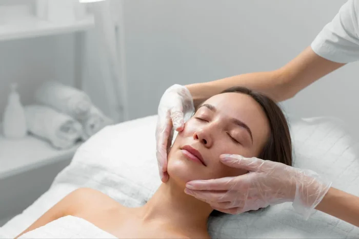 A woman lies on a spa bed with her eyes closed while a professional wearing gloves gently massages her face in a clean, bright treatment room.