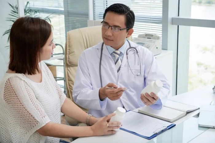 A doctor in a white coat sits at a desk, holding a pill bottle and talking to a female patient, who also holds a bottle. They are discussing medication in a bright medical office.