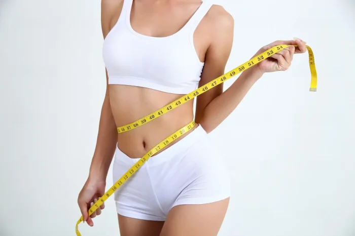 A woman in white shorts and a tank top measures her waist with a yellow measuring tape, standing against a plain white background.