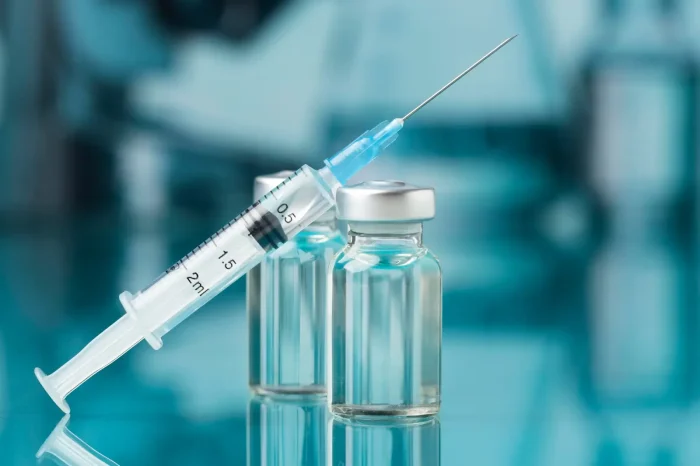 A syringe filled with clear liquid rests on two small glass vials with silver caps, set against a blue, blurred background.
