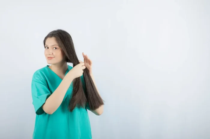 A woman in teal scrubs stands against a light background, holding and inspecting her long, dark hair with both hands, looking slightly to the side with a gentle smile.
