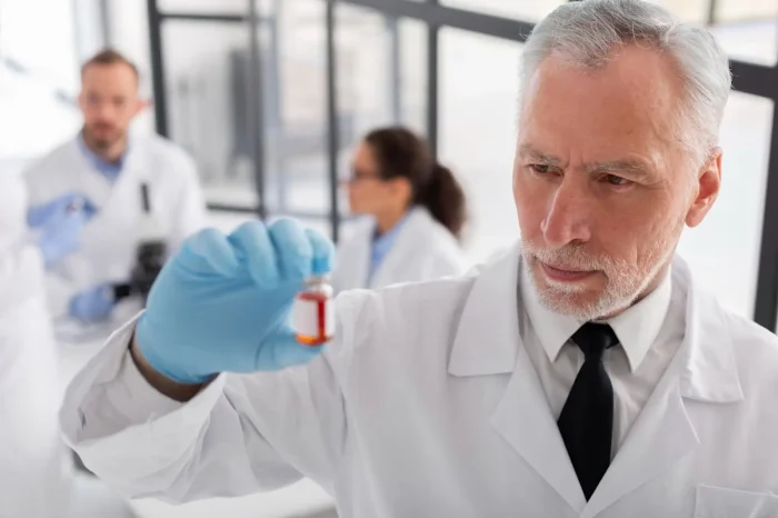 An older male scientist in a lab coat and gloves examines a vial with a red liquid, while two other scientists work in the background in a laboratory setting.