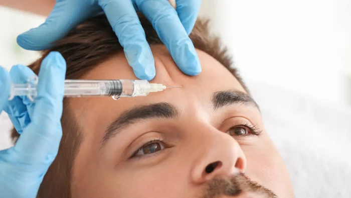 A close-up of a person receiving an injection in the forehead, administered by gloved hands, likely for a cosmetic procedure such as Botox.
