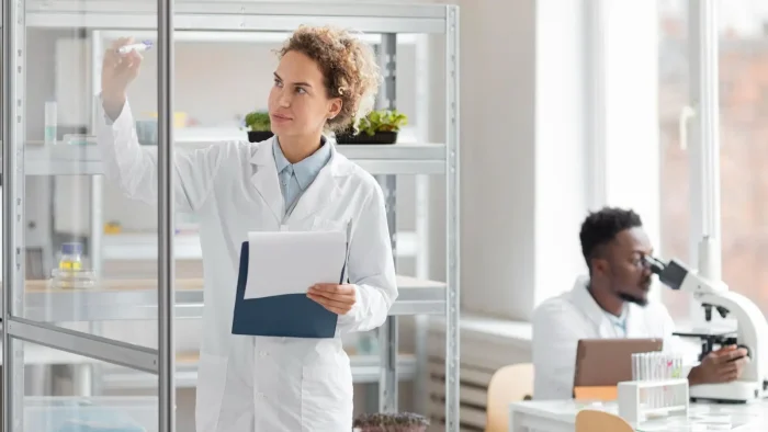 A woman in a lab coat writes on a transparent board while holding papers. In the background, a man in a lab coat looks through a microscope at a desk in a bright laboratory with plants and equipment.