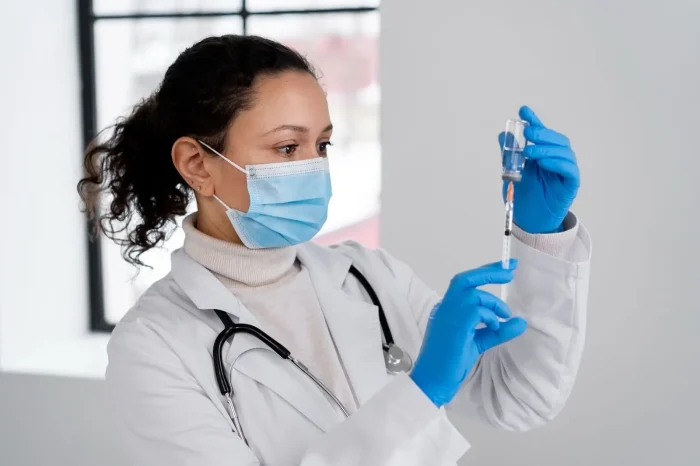 A healthcare worker wearing a lab coat, stethoscope, face mask, and blue gloves prepares a syringe with medication from a vial in a clinical setting.