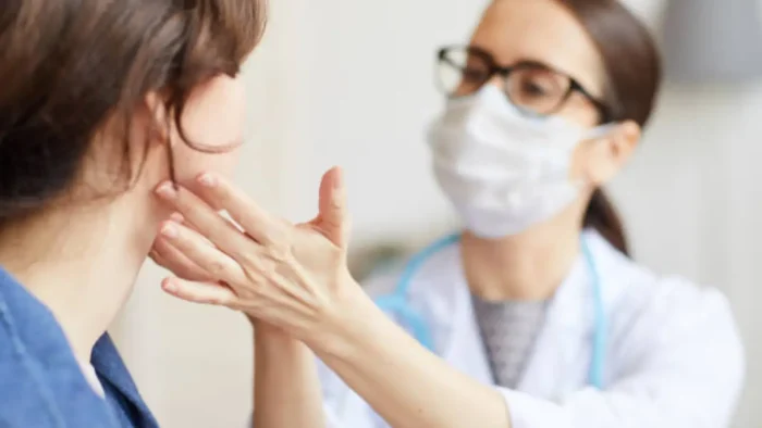 A doctor wearing a face mask and glasses examines a patient's neck. The doctor is gently pressing on the patient's neck area, possibly checking for swelling or abnormalities.
