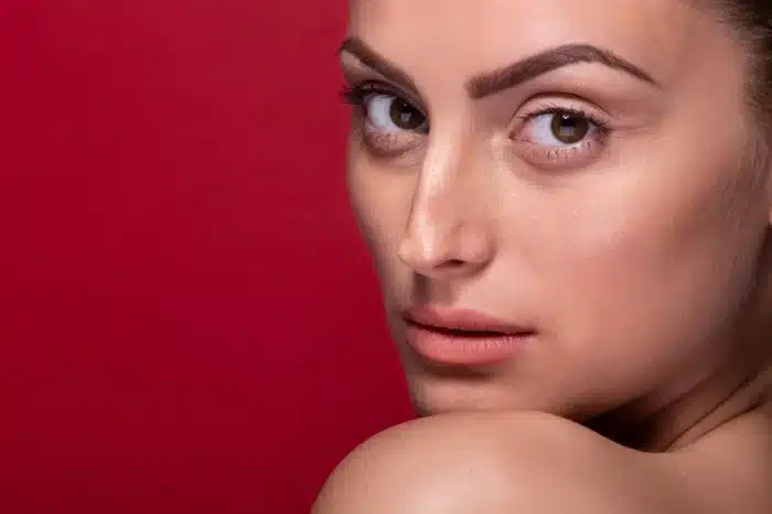 Close-up of a woman with smooth skin and defined eyebrows looking over her bare shoulder, set against a solid red background.