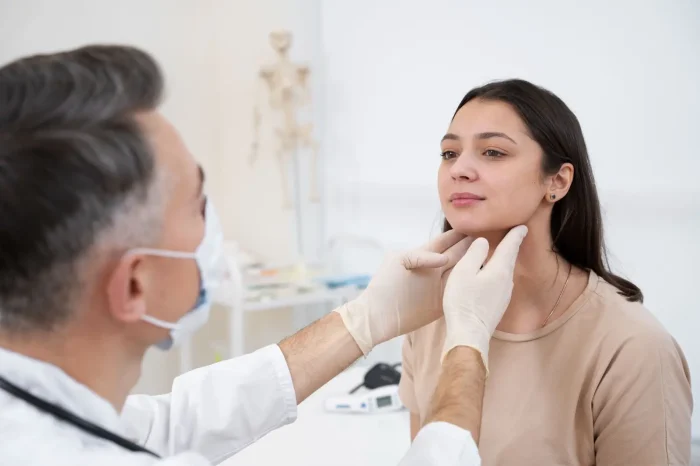A doctor wearing gloves and a mask examines a young woman's neck in a medical office. The woman looks calm, and medical equipment is visible in the background.