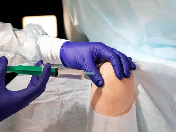 A medical professional wearing purple gloves prepares to inject a syringe into a patient's knee, which is draped with a sterile cloth in a clinical setting.