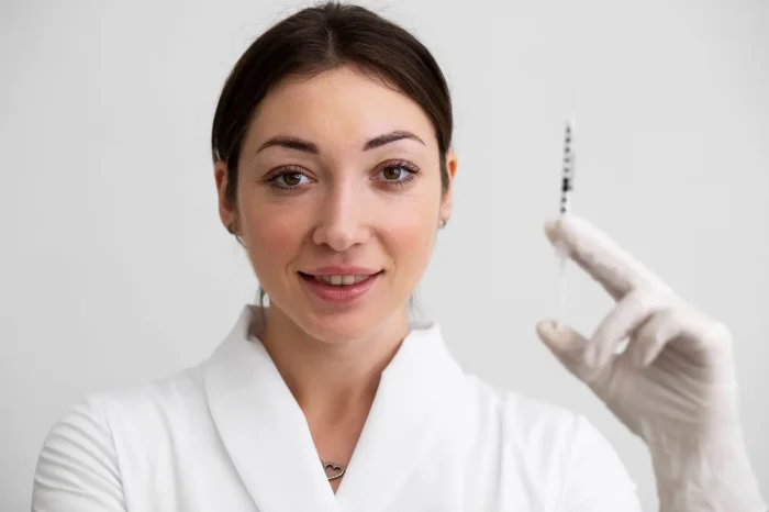 A woman in a white coat and gloves holds up a syringe, smiling slightly, against a plain light background.