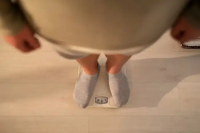 A person wearing gray socks stands on a white analog bathroom scale, viewed from above, on a light-colored wooden floor.
