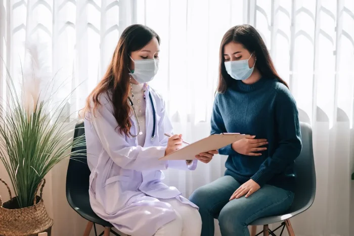 A doctor in a white coat and face mask sits with a clipboard, talking to a masked patient in a blue sweater who holds her stomach, in a bright room with large windows and a potted plant.