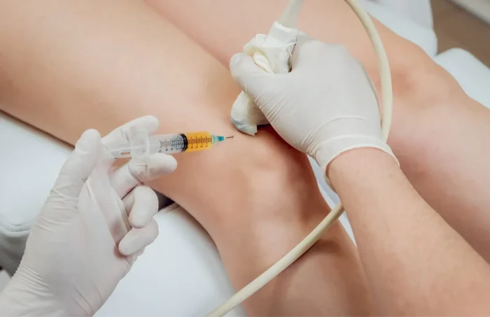 A healthcare professional wearing gloves uses an ultrasound device and syringe to perform an injection into a patient’s knee, likely for medical treatment or pain relief.