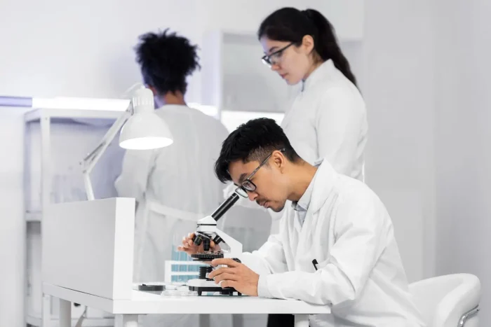 A male scientist in a white lab coat examines samples through a microscope at a lab desk, while two female colleagues work in the background, also wearing lab coats. The laboratory is bright and modern.
