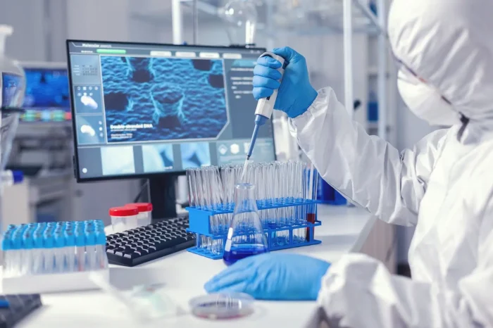 A scientist in protective gear uses a pipette to transfer blue liquid into test tubes in a laboratory, with a computer displaying scientific images in the background.