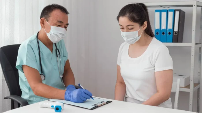 A doctor wearing scrubs, gloves, and a face mask discusses a medical form with a masked female patient in a white shirt. They are seated at a desk in a bright, organized office.