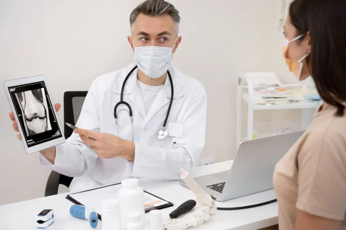 A doctor wearing a mask shows and explains a knee X-ray on a tablet to a patient, who is also masked, in a medical office with a laptop, spine model, and medical supplies on the desk.
