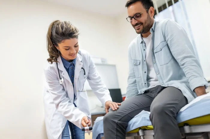 A female doctor checks a male patient's knee reflex with a reflex hammer as he sits on an examination table, smiling, in a medical office.