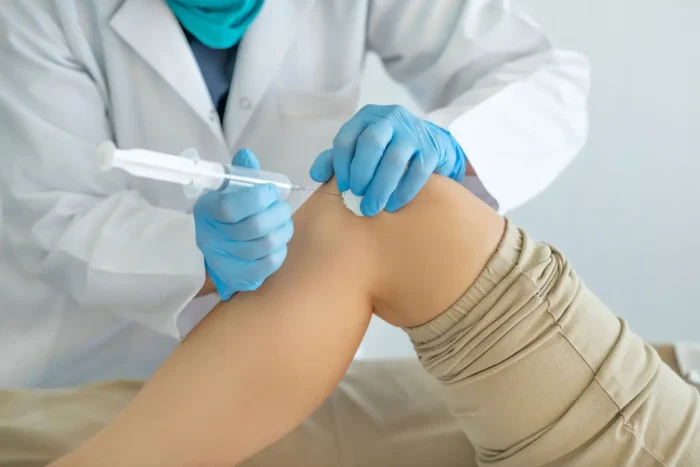 A medical professional wearing gloves and a lab coat injects a needle into a patient's knee, suggesting a medical procedure or treatment for knee pain or injury.