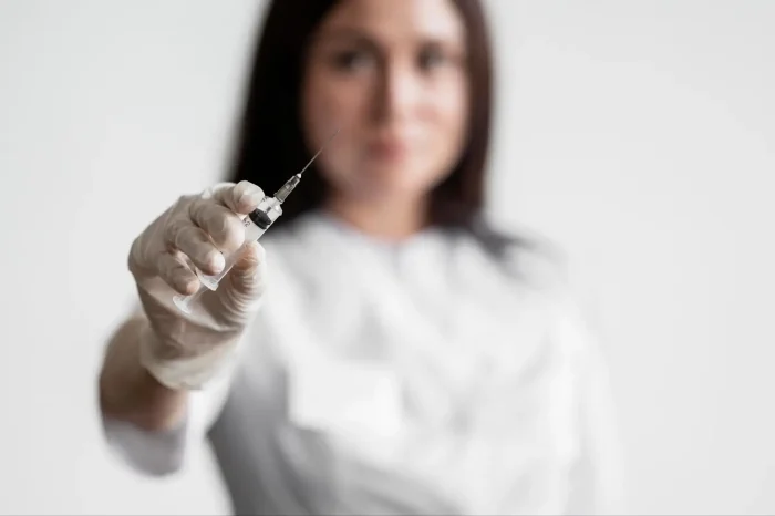 A person wearing white gloves and a lab coat holds a syringe towards the camera, with the syringe in focus and the person blurred in the background.