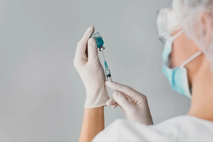 A healthcare professional wearing a mask, face shield, and gloves prepares a syringe with blue liquid from a vial, set against a light grey background.
