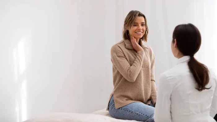 A woman sits on an examination table, smiling and touching her neck, while talking to a healthcare professional in a white coat, in a bright, minimalistic room.