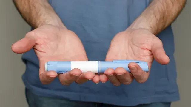 A person wearing a blue shirt holds an insulin pen with both hands, palms up, against a neutral background.