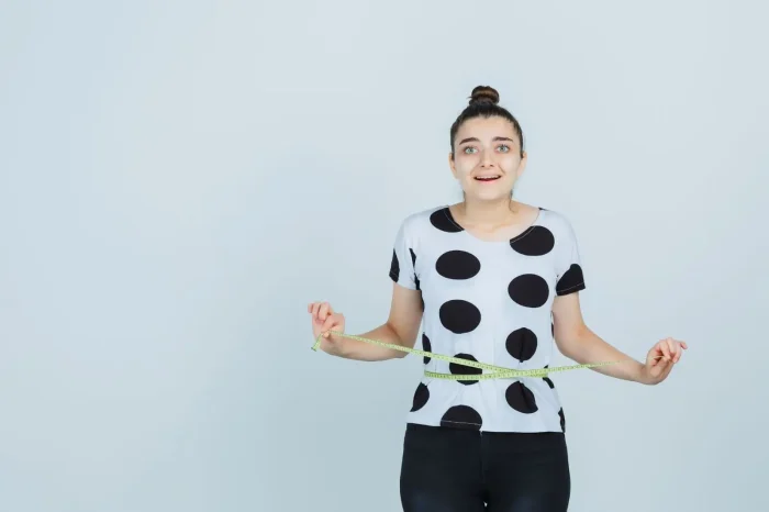 A young woman with a surprised expression holds a measuring tape around her waist. She is wearing a white shirt with large black polka dots and black pants, standing against a plain light gray background.