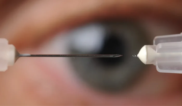 A close-up of a needle with a droplet at its tip, positioned in front of a human eye, which is out of focus in the background.