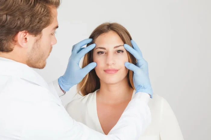 A doctor wearing blue gloves examines a woman's face by gently touching her forehead and cheekbones. The woman, with brown hair and a neutral expression, looks directly at the camera. Both are dressed in white.