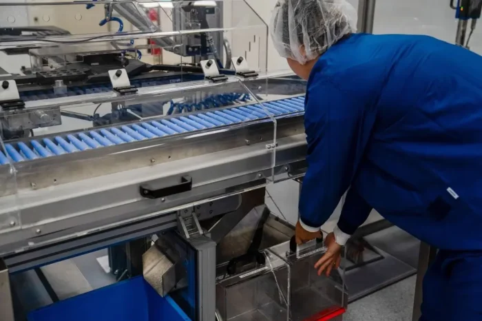 A worker wearing a blue lab coat and hairnet operates a machine in a factory, collecting items from a conveyor belt into a clear plastic container.