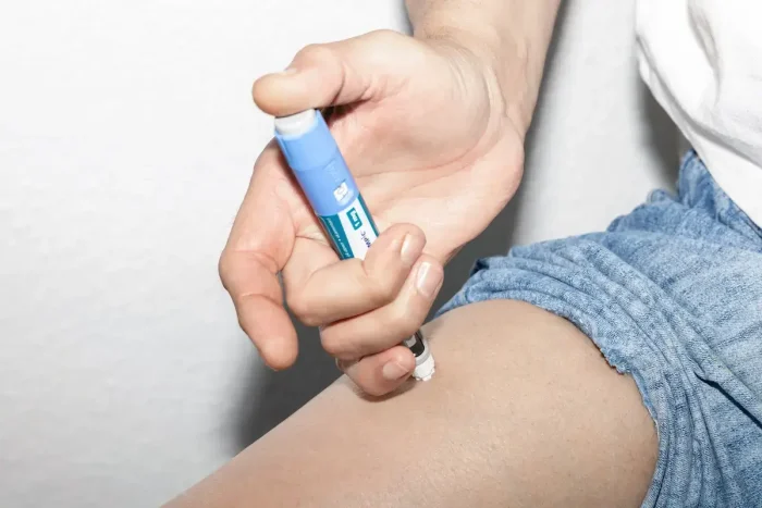 A person injects medication into their thigh using an insulin pen. They are wearing shorts, and the focus is on their hand and the injection site.