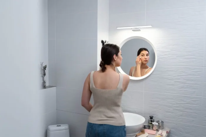 A woman stands in front of a bathroom mirror, applying makeup to her face. The counter in front of her has various cosmetic products and toiletries. The bathroom is modern and well-lit.