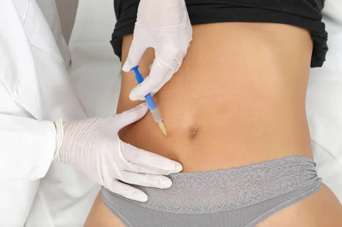 A person wearing white gloves injects a syringe into the lower abdomen of a patient lying down, who is dressed in a black top and gray lace underwear.