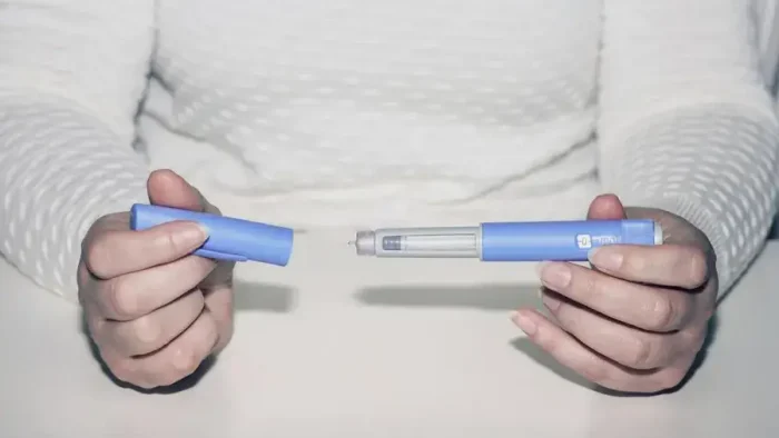 A person wearing a white textured sweater holds an insulin pen, with one hand removing the cap, revealing the needle for injection.