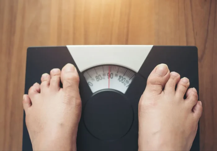 Close-up of bare feet standing on a mechanical bathroom scale, displaying a weight of approximately 95 kilograms, on a wooden floor background.