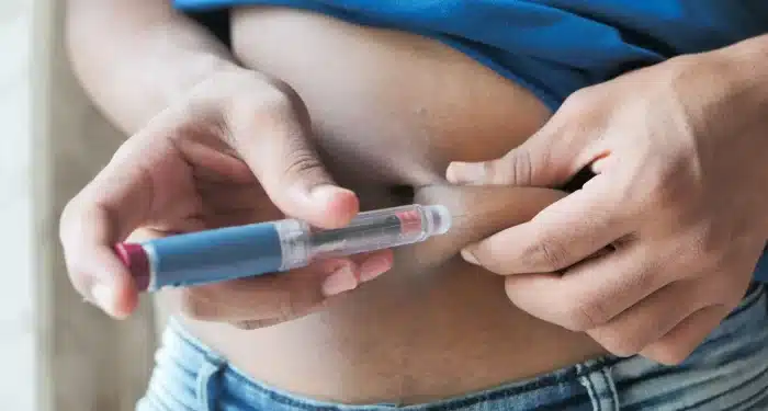 Close-up of a person injecting medication into their abdomen with an insulin pen, holding the skin with one hand and the injector in the other. The person is wearing jeans and a blue shirt.