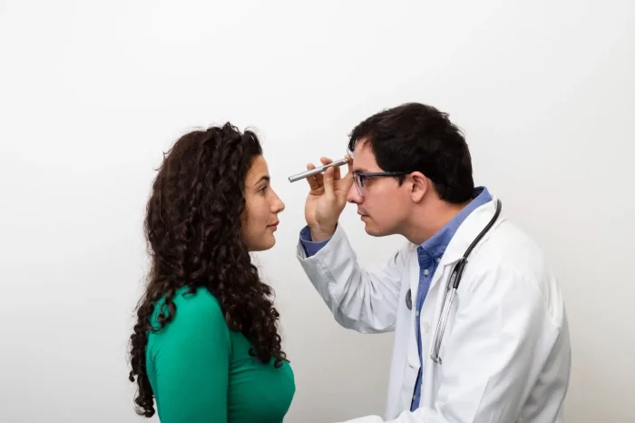A doctor in a white coat examines a woman's eyes using a small flashlight. The woman, wearing a green top, faces the doctor against a plain white background.