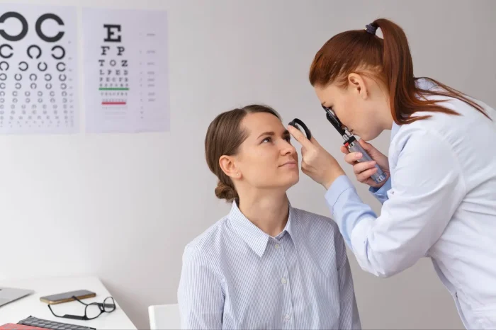 An eye doctor examines a patient's eyes with an ophthalmoscope in a clinic; eye charts are visible on the wall in the background.
