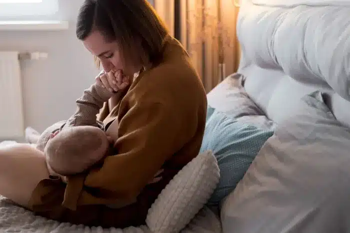 A mother sits on a bed and breastfeeds her baby, gently holding and kissing the baby’s hand. The room is softly lit, creating a warm and intimate atmosphere. Pillows and blankets surround them.