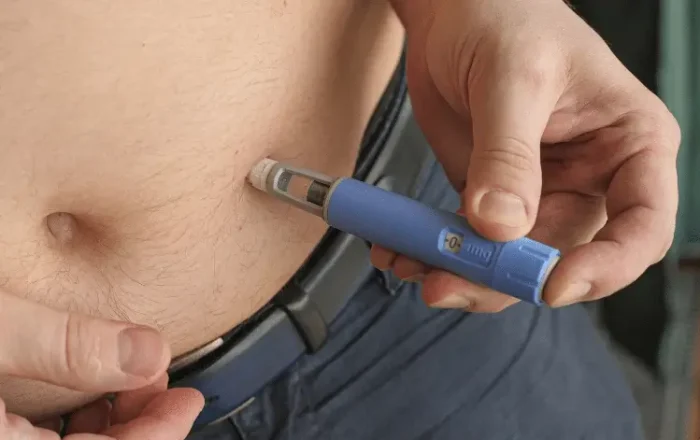 A person injects medication into their abdomen using a blue injector pen, with one hand holding the pen and the other pulling up their shirt.