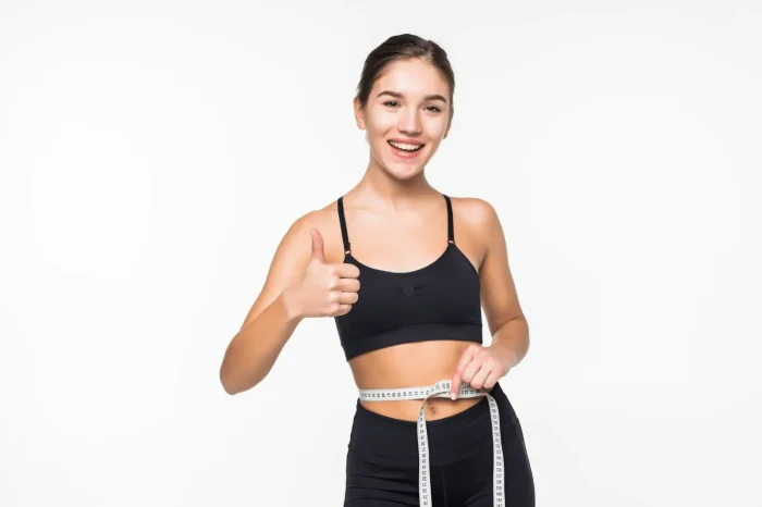 A young woman in black sportswear smiles, holding a measuring tape around her waist and giving a thumbs-up gesture, standing against a plain white background.