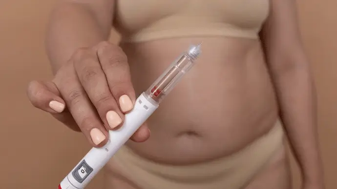 A person wearing beige underwear holds a medication injection pen toward the camera, with their midsection and navel visible in the background.