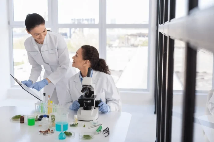 Two female scientists in lab coats and gloves work in a bright laboratory. One is standing with a clipboard, while the other sits at a microscope. Test tubes and plant samples are on the table. They are smiling at each other.