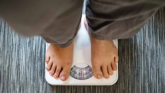A person wearing gray pants stands on a white analog bathroom scale, which is placed on a textured gray surface. The scale displays a weight measurement.