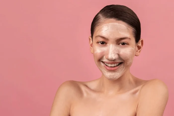 A smiling person with slicked-back hair stands against a pink background, wearing a white facial mask on their face and shoulders bare.